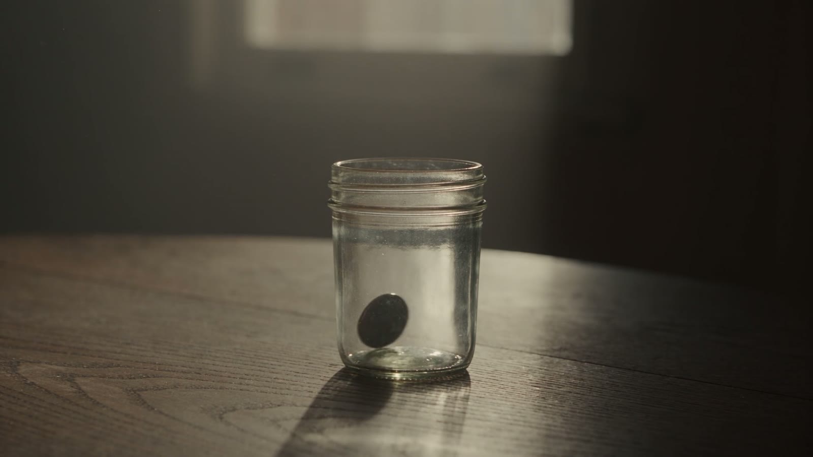 A single coin drops into an empty glass jar on a bare desk.